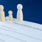 Printed child support agreement with man, woman and child wooden figures in a conceptual image for financial child support. Over blue background.