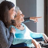 Joyful woman taking care of elderly mother. Young woman hugging senior lady and showing something out of window. Positive emotions concept.